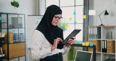 Güzel, güler yüzlü, kendinden emin, geleneksel türbanlı Müslüman bir kadın modern ofiste kameraya yan bakıyor ve tablet bilgisayar kullanıyor.