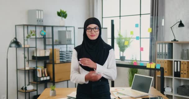 Atractiva sonriente mujer musulmana exitosa con confianza en el hijab de pie en la oficina moderna y mirando a la cámara con los brazos cruzados, vista frontal, cámara lenta — Vídeo de stock