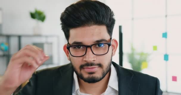 Aantrekkelijke glimlach succesvolle jonge bebaarde office manager neemt zijn bril af terwijl poseren op camera op de achtergrond van modern kantoor, close-up — Stockvideo