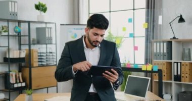 Yakışıklı, çalışkan, zeki, sakallı, takım elbiseli genç bir işadamı kameranın önünde duruyor ve modern ofiste tablet bilgisayarı üzerinde çalışıyor.