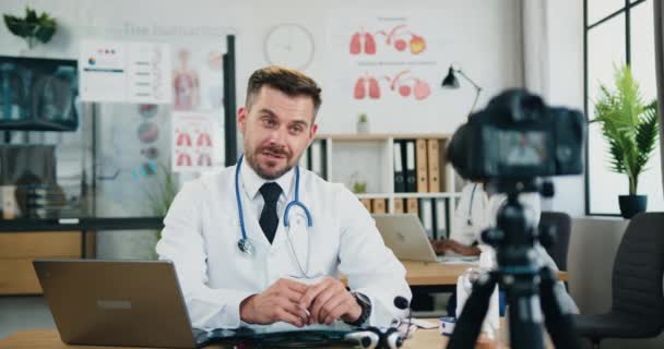 Atractivo médico barbudo con experiencia y confianza sentado frente a la cámara en su propio consultorio médico y grabar video para su audiencia en Internet — Vídeos de Stock