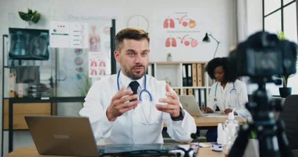 Provável confiante inteligente médico barbudo qualificado sentado em seu local de trabalho no consultório médico e gravação de vídeo na câmera para o público da internet — Vídeo de Stock