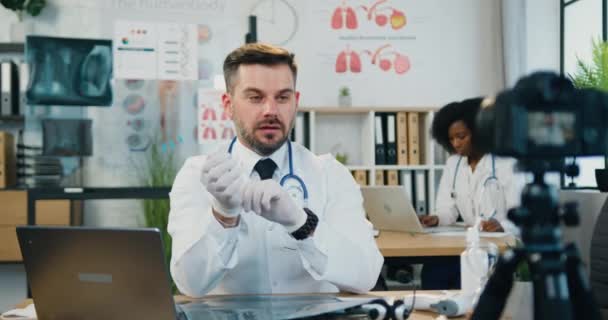 Medisch blogconcept waarbij aantrekkelijke positief gekwalificeerde volwassen bebaarde medic video opneemt op camera voor medisch kanaal en laat zien hoe medische handschoenen worden aangetrokken — Stockvideo