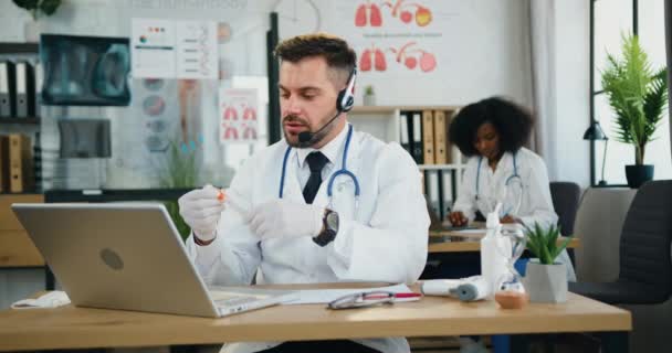 Крупным планом симпатичного уверенного умного бородатого врача в наушниках, сидящего перед ноутбуком во время видеочата с пациентом и объясняющего, как использовать шприц для инъекций — стоковое видео