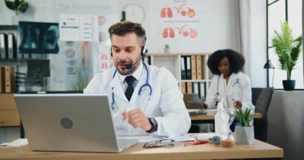 Retrato de simpático médico barbudo experto positivo en uniforme y guantes en auriculares que celebran una reunión de vídeo con colegas en el consultorio médico en el fondo de su negro de trabajo — Vídeo de stock