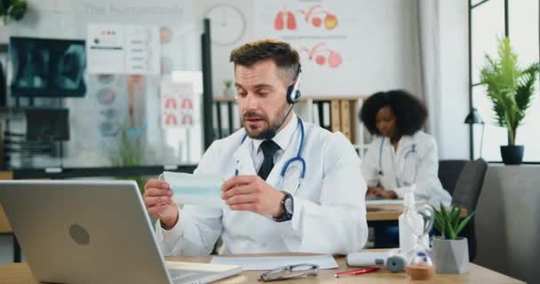 Likable collected skilled professional bearded doctor in wireless headset telling to online audience about the need to wear protective mask in time of pandemic covid-19 — Stock Video