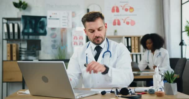 Portrét dobře vypadajícího seriózního úspěšného zkušeného vousatého lékaře, který si nasazuje brýle při práci v laptopu v lékařské laboratoři spolu s černošskou asistentkou — Stock video