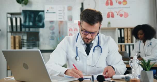 Sympatický kvalifikovaný vousatý doktor v brýlích, který pracuje s lékařskými papíry a píše na počítači na klinice poblíž své mladé kolegyně s černou pletí — Stock video