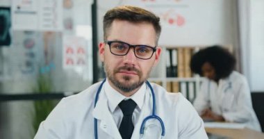 Çekici, olumlu, başarılı, zeki, sakallı doktor kameraya bakarken gözlüğünü çıkarıyor. Siyah tenli bayan meslektaşının arka planında samimi bir gülümsemeyle.