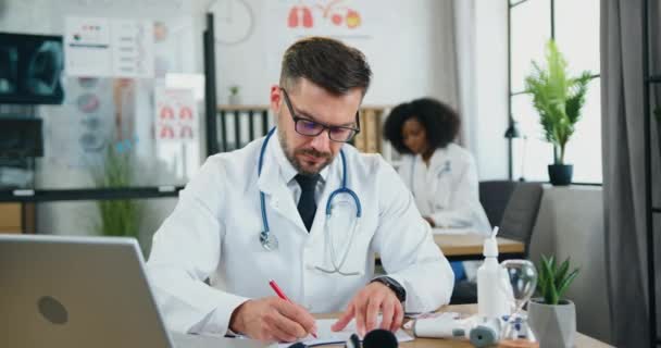 Attractive concentrated confident experienced bearded doctor in glasses writing notes into patients medical card and looking at camera on working black-skinned female assistant background — Stock Video