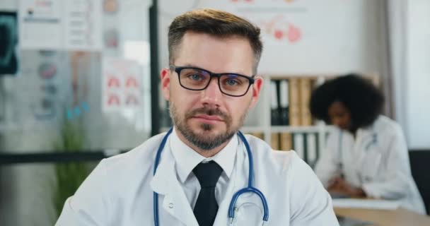 Aantrekkelijk positief tevreden succesvolle slimme bebaarde arts die zijn bril afdoet wanneer hij met oprechte glimlach in de camera kijkt op de achtergrond van een zwarte vrouwelijke collega — Stockvideo