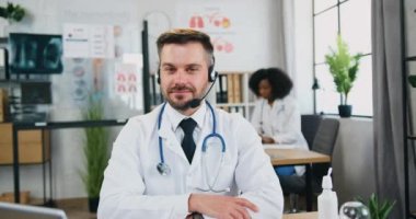 Çekici, gülümseyen, zeki, sakallı, kulaklıklı doktor. Tıp laboratuarında kameranın önünde, siyah tenli bayan meslektaşının arka planında oturuyor.