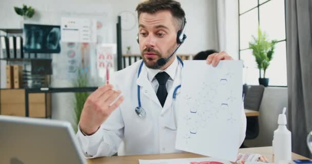 Atractivo profesional de confianza con experiencia médico barbudo en auriculares celebración de chat de vídeo y la explicación de la fórmula química para la audiencia a través de ordenador portátil — Vídeo de stock