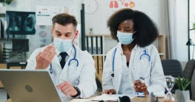 Çekici, kendine güvenen, koruyucu maskeli erkek ve kadın melez yarış doktorları dizüstü bilgisayarda online seminer düzenliyor ve covid-19 döneminde maske takmaları gerektiğini anlatıyor.