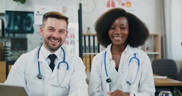 Atractivo amigable sonriendo exitosos médicos de raza mixta calificados mirando a la cámara con expresiones de la cara feliz y gestos pulgares hacia arriba — Vídeos de Stock