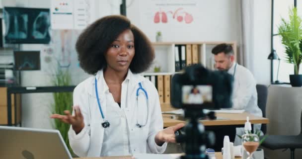 Beautiful positive confident skilled female black-skinned doctor recording videovlog on camera for online audience from medical room on the background of hardworking on laptop male colleague in the — Stock Video