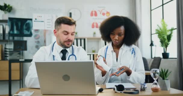 Concepto de medicina donde atractivo positivo experimentado multirracial dos trabajadores médicos celebración de seminario en línea a través de la computadora portátil y explicar cómo utilizar termómetro infrarrojo sin contacto — Vídeo de stock