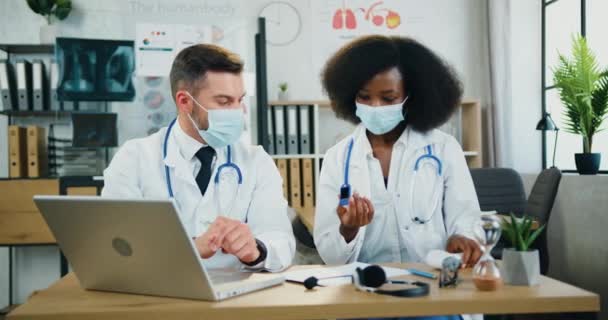 Likvidovatelné sebevědomé kvalifikované dva smíšené rasy lékaři v ochranných maskách vzdáleně podporující svého pacienta během videohovoru na notebooku z lékařské kanceláře a vysvětlující, jak používat infračervený teploměr — Stock video