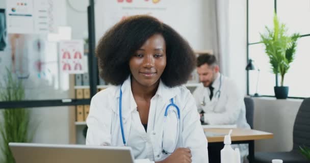 Atractiva sonriente experimentada trabajadora médica de piel negra sentada frente a la cámara en el consultorio médico y mirando con expresión de cara feliz en el fondo del hombre barbudo trabajador — Vídeo de stock