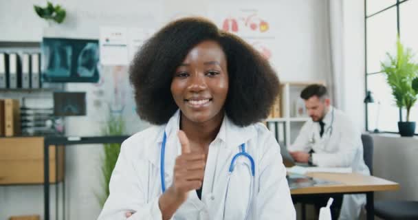 Attrayant souriant satisfait qualifié afro-américain médecin en uniforme médical avec stéthoscope posant à la caméra dans le bureau médical moderne et montrant pouce vers le haut — Video