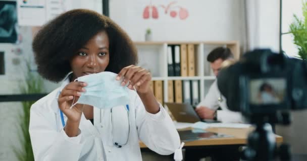 Primo piano di bello positivo fiducioso esperto afro-americano medico femminile che streaming sulla macchina fotografica dallo studio medico e insegnare agli spettatori come corretta indossare maschera protettiva — Video Stock