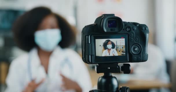 Telemedicine concept where cute confident professional qualified black-skinned nurse in medical mask recording video for internet audience on camera and explaining about the need to wear masks in — Stock Video