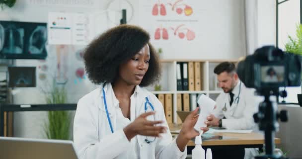 Beautiful positive qualified smart black-skinned female doctor recording video on camera in modern medical office and explaining how to use infrared non-contact thermometer for internet viewers — Stock Video