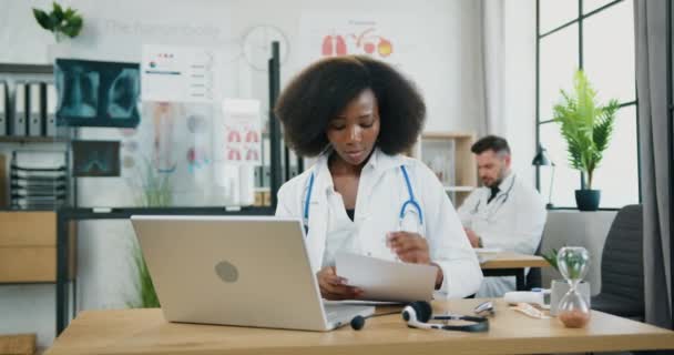 Bella positiva fiduciosa esperto medico donna dalla pelle nera in uniforme bianca prendere appunti in documenti durante la riunione video sul computer portatile nel moderno studio medico — Video Stock