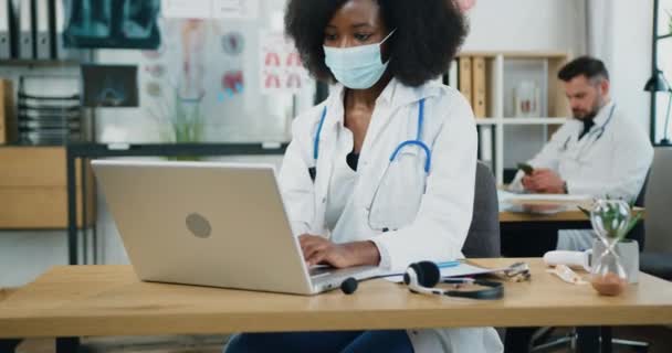 相貌俊俏、技术精湛的黑皮肤女医生，戴着面罩，坐在笔记本电脑前的桌子旁，在留胡子男性同事的背景下打字，医学概念 — 图库视频影像