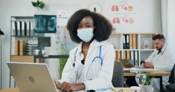 Söt säker högkvalificerad svarthyade kvinna-läkare i medicinsk ansiktsmask tittar in i kameran under arbete på datorn i modern klinik kontor nära skäggiga manliga kollega — Stockvideo