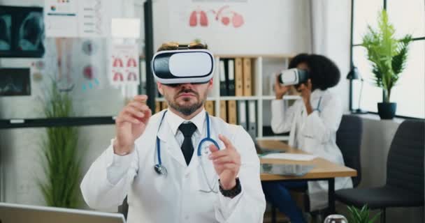 Atractivo médico barbudo cualificado serio y confiado y su compañera de ayuda afroamericana trabajando juntos en gafas de realidad aumentada en el laboratorio médico moderno — Vídeo de stock