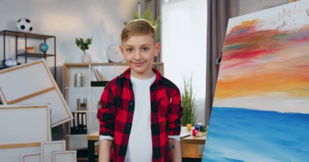 Concetto di arti in cui carino sorridente ragazzo di 10 anni in posa sulla macchina fotografica in laboratorio di pittura e mostrando molti pennelli in macchina fotografica, rallentatore — Video Stock