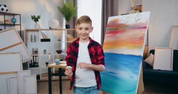 Närbild av härlig glad leende skicklig blond skolpojke som poserar på kamera med korsade armar med borste under teckning bild i hemmet verkstad — Stockvideo