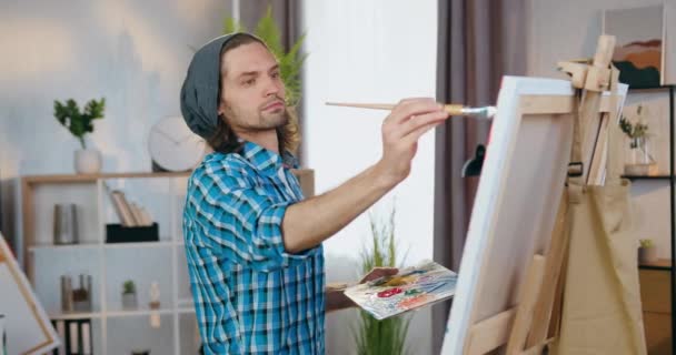 Dobře vypadající sebevědomý kreativní mladý kluk-malíř kreslení obrázek s štětcem na plátně připevněné k stojanu v krásně zdobené domácí dílny ve dne — Stock video