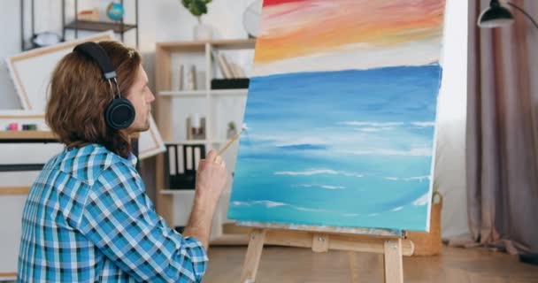 Attractive confident skilful bearded young man-artist drawing picture and enjoying music in headphones in own home studio — Stock Video