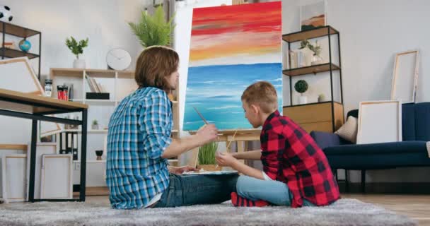 Zręczny, pewny siebie, brodaty ojciec-artysta i jego 10-letni syn rysują razem obraz na płótnie z pędzlami w domu — Wideo stockowe