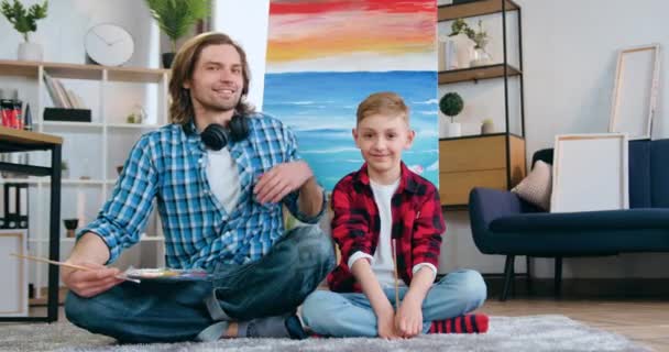 Gros plan portrait de beau sourire heureux homme barbu talentueux et son fils qui assis sur le sol près de l'image dessinée sur toile et regardant dans la caméra avec des pinceaux — Video