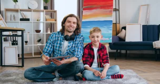 Przystojny uśmiechnięty brodaty mężczyzna i uroczy chłopiec obaj artyści siedzący przed kamerą w pobliżu malowanego obrazu na płótnie i trzymający pędzle i paletę z farbami — Wideo stockowe