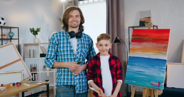 Gros plan de beau sourire talentueux homme-artiste moderne et son petit garçon étudiant qui pose sur la caméra dans un studio d'art près de l'image dessinée — Video