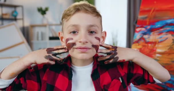 Милий усміхнений грайливий хлопчик з руками в фарбах малює лінії на обличчі в домашній художній студії, безтурботний концепт дитинства — стокове відео