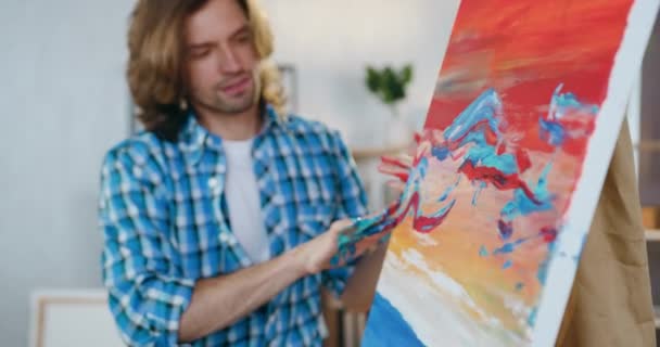 Goed uitziende zelfverzekerde jonge bebaarde schilder die verf met de vingers op groot doek aanbrengt tijdens het maken van een abstract beeld in zijn atelier — Stockvideo