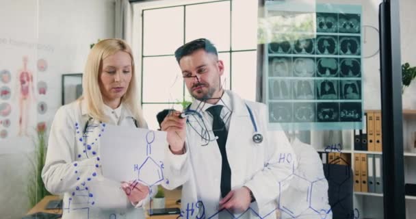 Медицинская и научная концепция, где привлекательные уверенные высококвалифицированные взрослые врачи стоят у стеклянной стены в медицинской лаборатории и пишут химическую формулу на этой стене — стоковое видео