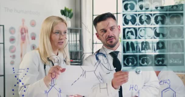 Вид спереду позитивний досвідчені дорослі лікарі чоловічої та жіночої статі, які стоять біля скляної стіни з рентгенівським зображенням пацієнтів і розмовляють один з одним про діагноз — стокове відео