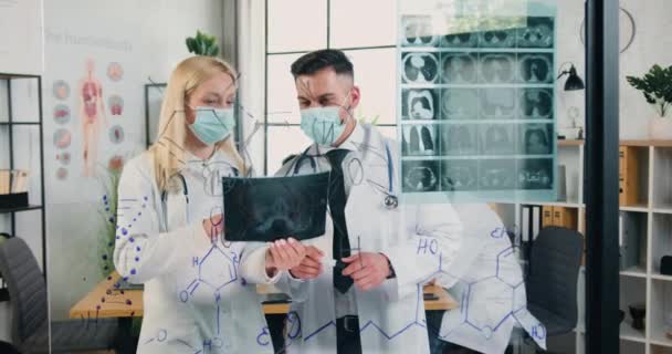 Medycyna w czasie covid-19, gdzie dobrze wyglądająca pewna siebie profesjonalna dorosła kobieta i mężczyzna współpracownicy medyczni w maskach ochronnych pracujących ze skanerem rentgenowskim przymocowanym do szklanej ściany w laboratorium medycznym — Wideo stockowe