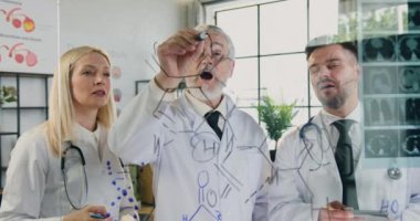 Tıbbi laboratuvarda cam duvarın yanında dikilen ve olgun sakallı ana doktorun kimyasal formül yazmasını izleyen farklı yaşlarda yoğun deneyimli tıp meslektaşları.