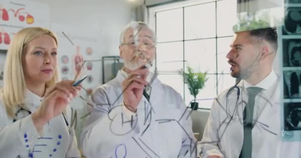 Lavoro in laboratorio ospedaliero dove attraente intelligente qualificato diverse età colleghi medici in piedi dietro parete di vetro e analizzando formula chimica scritta su questo muro — Video Stock