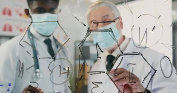 Close view through glass wall on good-looking successful experienced different ages mixed race doctors in sterile medical masks which discussing chemical formula drawn on this wall — Stock Video