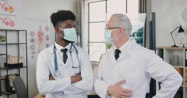 Portret van goed uitziende zelfverzekerde bekwame verschillende leeftijden gemengd ras artsen in beschermende maskers en witte jassen die op zoek naar camera in moderne kliniek kantoor — Stockvideo
