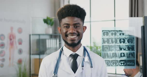 Primo piano di bello sorridente intelligente di successo qualificato giovane medico dalla pelle nera in cappotto bianco con stetoscopio in piedi davanti alla fotocamera in laboratorio clinica con espressione del viso soddisfatta — Video Stock