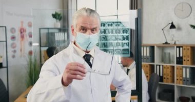 Kendine güvenen, yetenekli, gri saçlı, maske takan, modern tıp ofisinde kameraya poz veren, yetişkin erkek doktoru yakından çek.